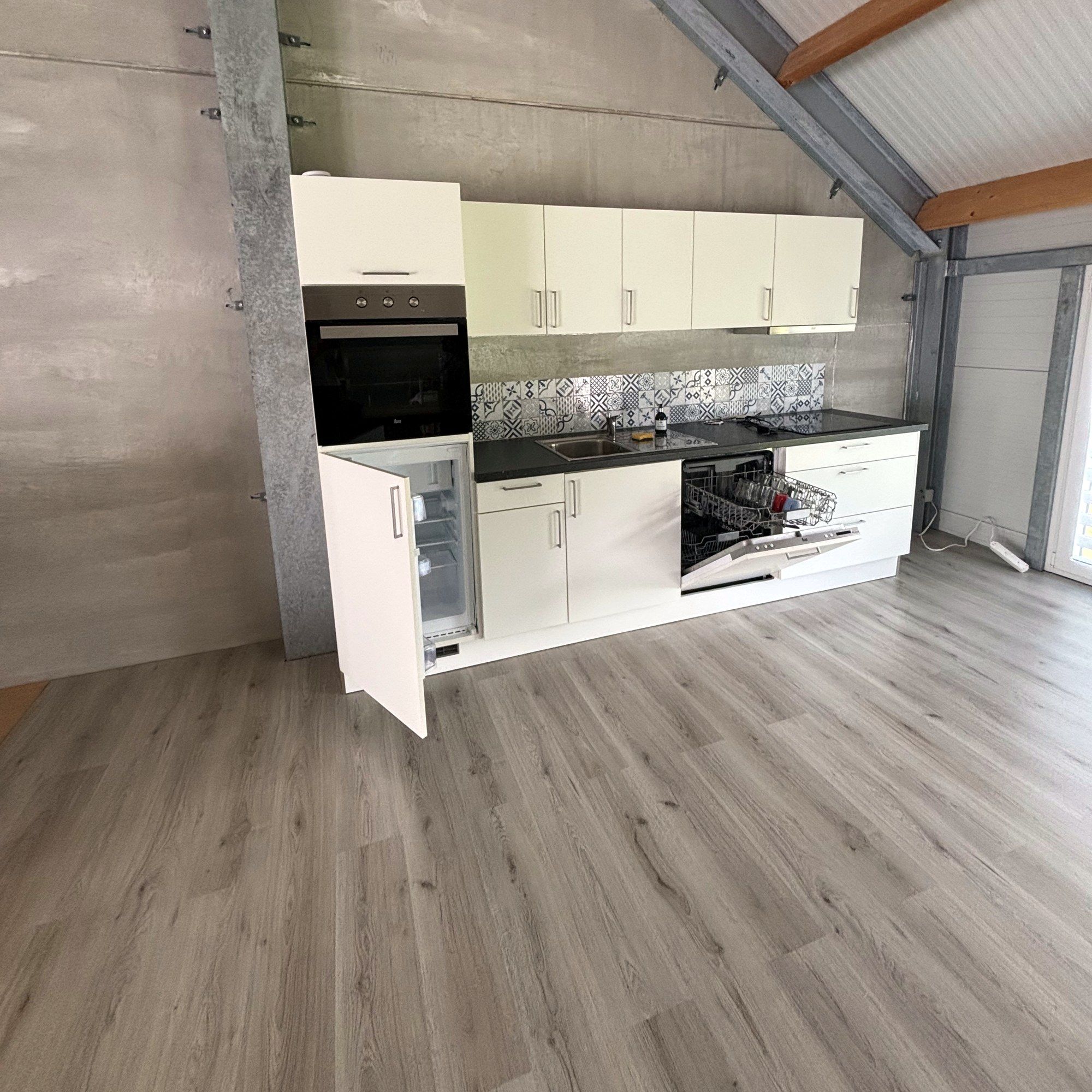 La cuisine linéaire existante, blanche avec plan de travail sombre, le long du mur sous la pente du toit.
